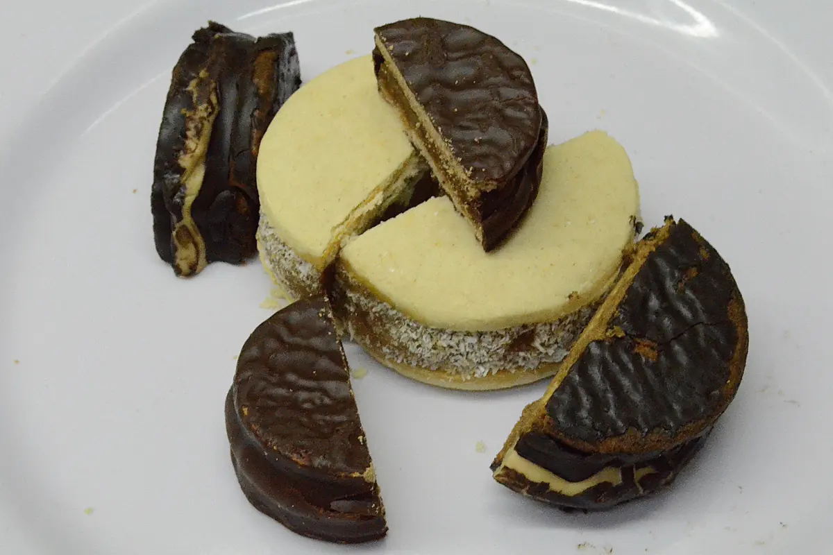 Argentinské alfajores na bílém talíři, v čokoládě a bílé glazuře, jeden rozkrojený, aby ukázal náplň
