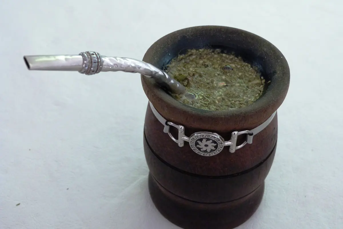 Uma cuia de mate de madeira com rebordo guarnecido a prata, cheia de erva-mate e uma bombilla de prata