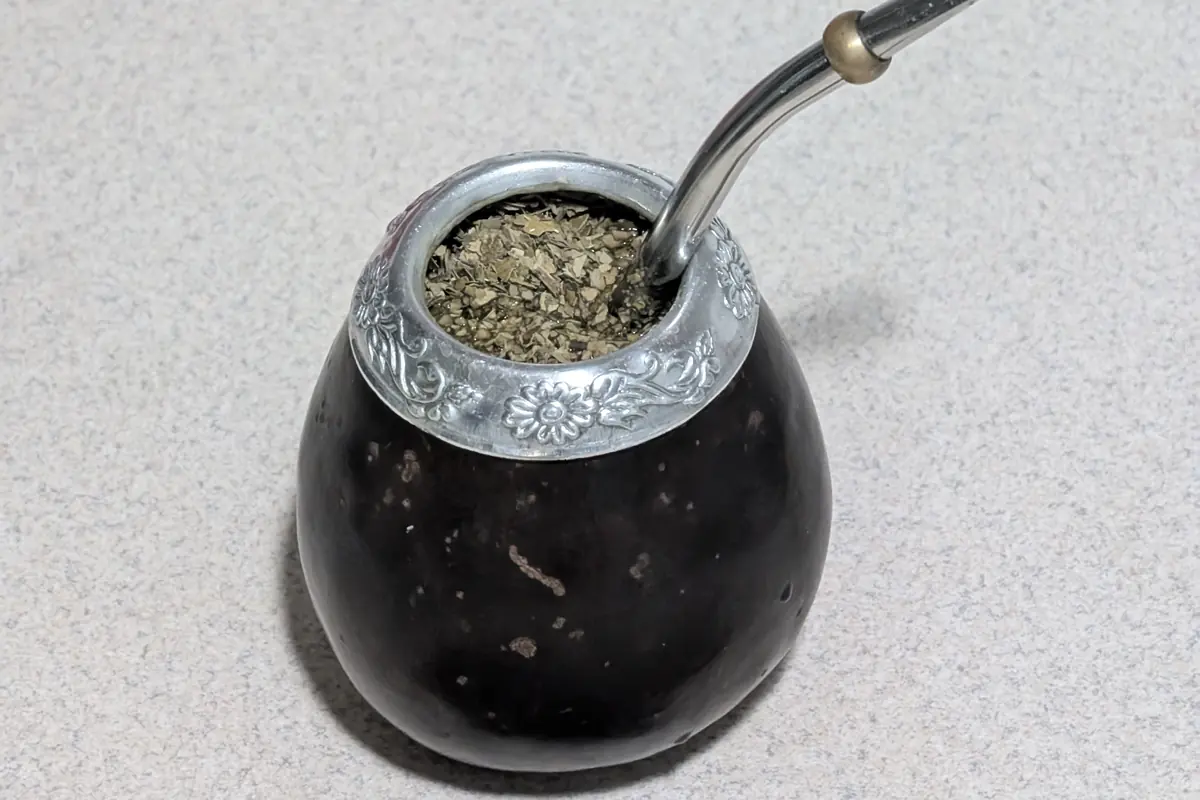 Černá tykev na maté se stříbrným okrajem, naplněná yerba maté a zahnutou kovovou bombillou, vedle dřevěné lžičky