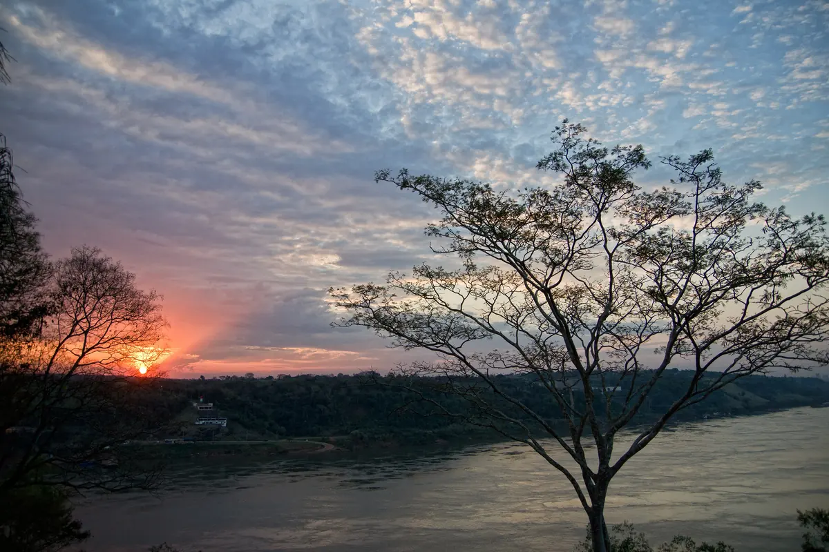 Západ slunce nad řekou u Hito Tres Fronteras v Puerto Iguazú, s holým stromem proti pruhovanému nebi